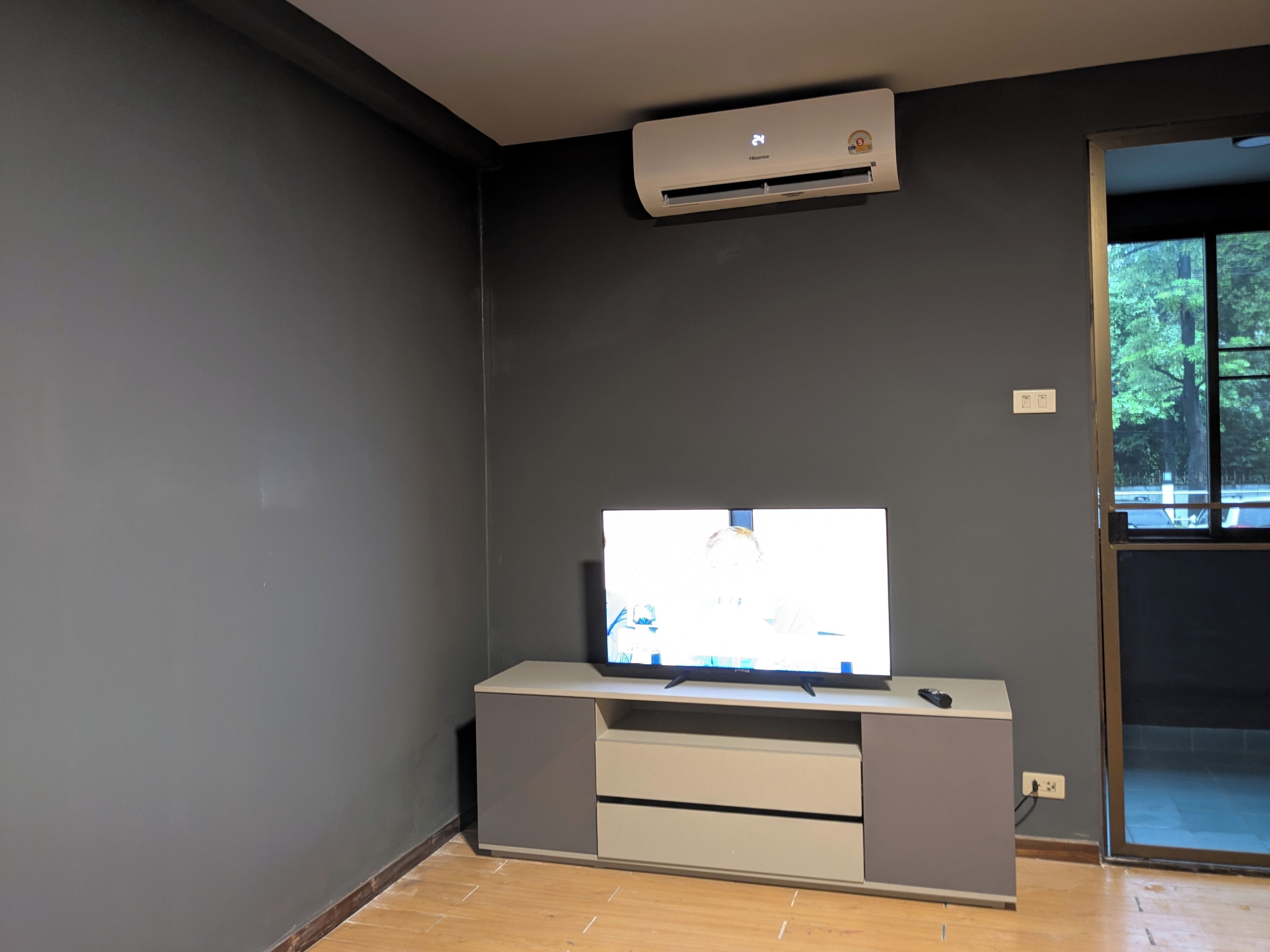 Habitación con televisor de pantalla plana sobre un mueble, equipo de aire acondicionado en la pared y ventana con vista a árboles.