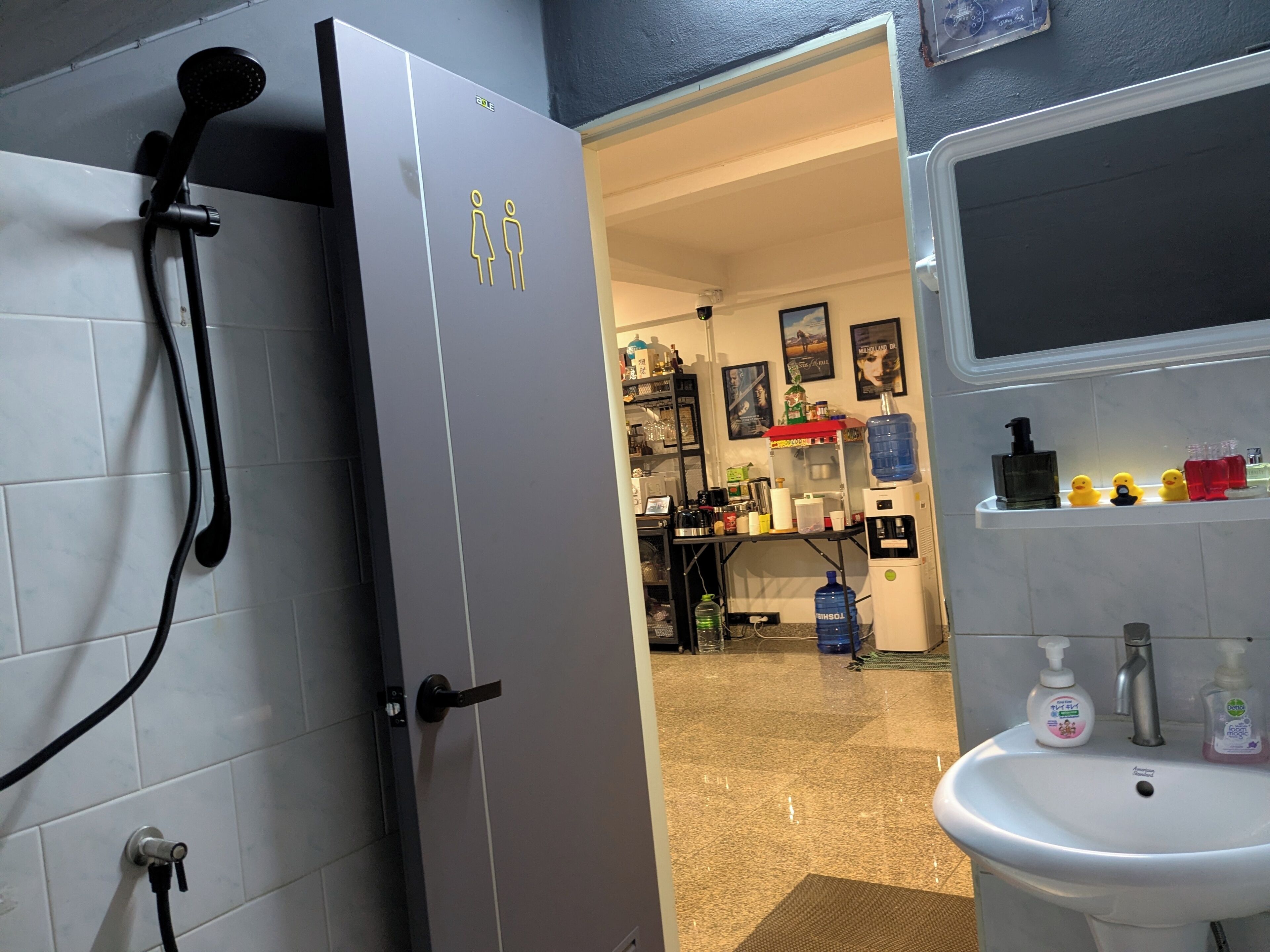 A bathroom with a shower, sink, and a door leading to a room with shelves and a coffee machine.