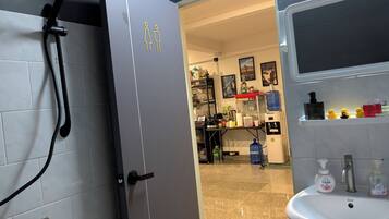 A bathroom with a shower, sink, and a door leading to a room with shelves and a coffee machine.