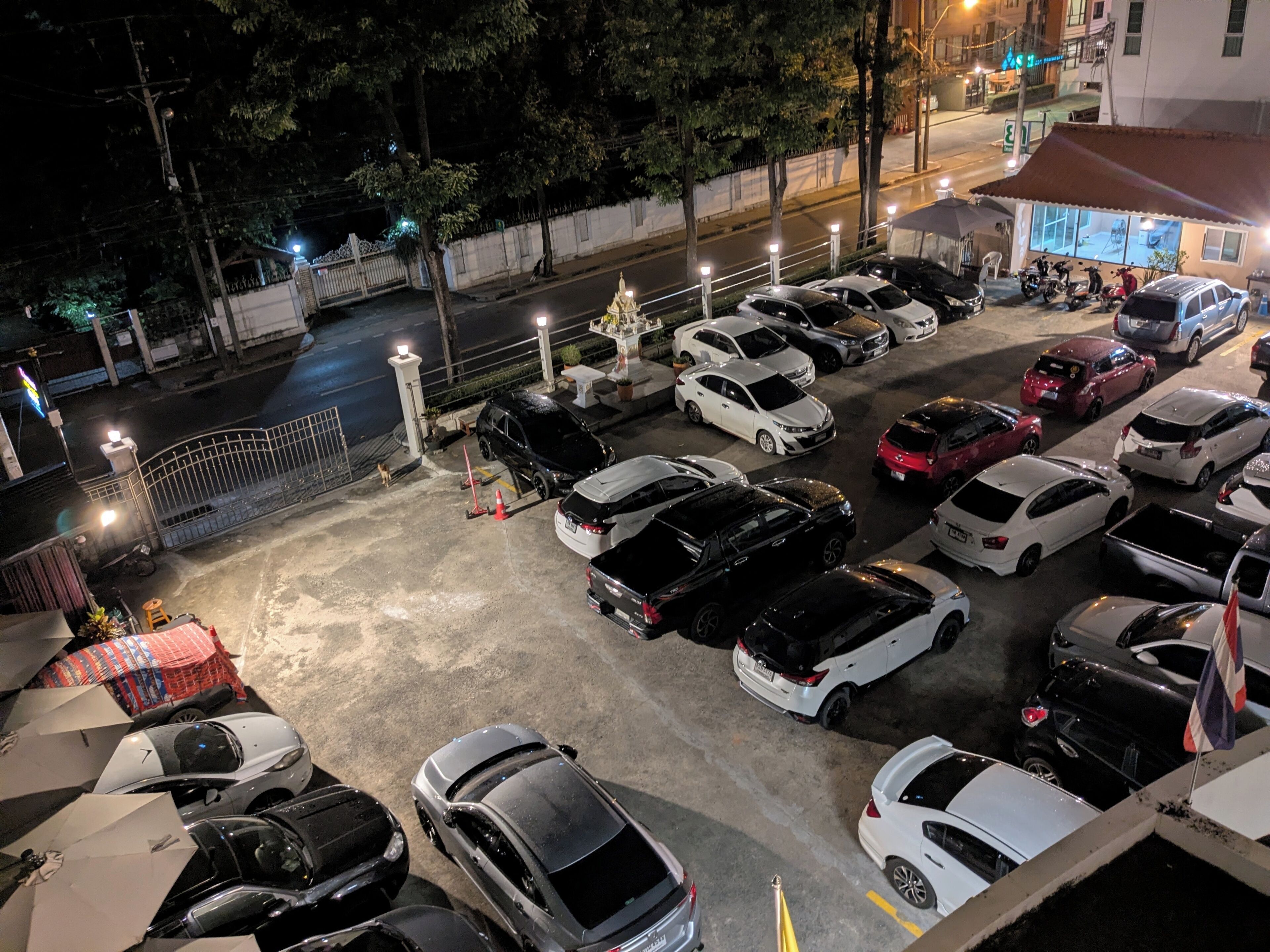 A parking lot with multiple cars and motorcycles, a gated entrance, and a building with illuminated windows in the background.