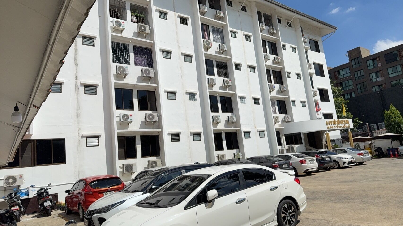 A multi-story building with air conditioning units on each floor, a parking lot with several cars, and a sign with Chinese characters.