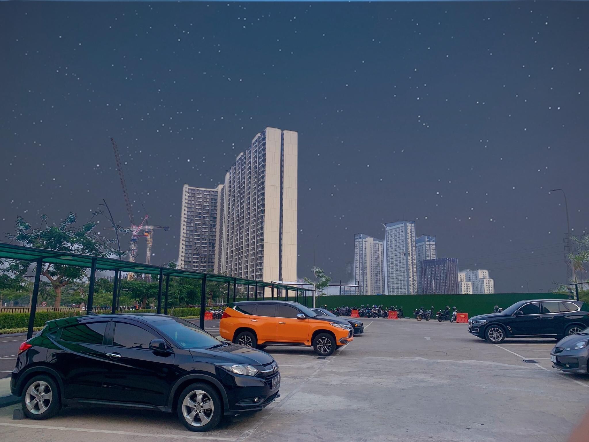 A parking lot with several cars, including a black SUV and an orange SUV, and a green fence in the foreground.