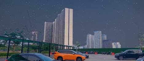 Un parking avec plusieurs voitures, dont un SUV noir et un SUV orange, et une clôture verte au premier plan.