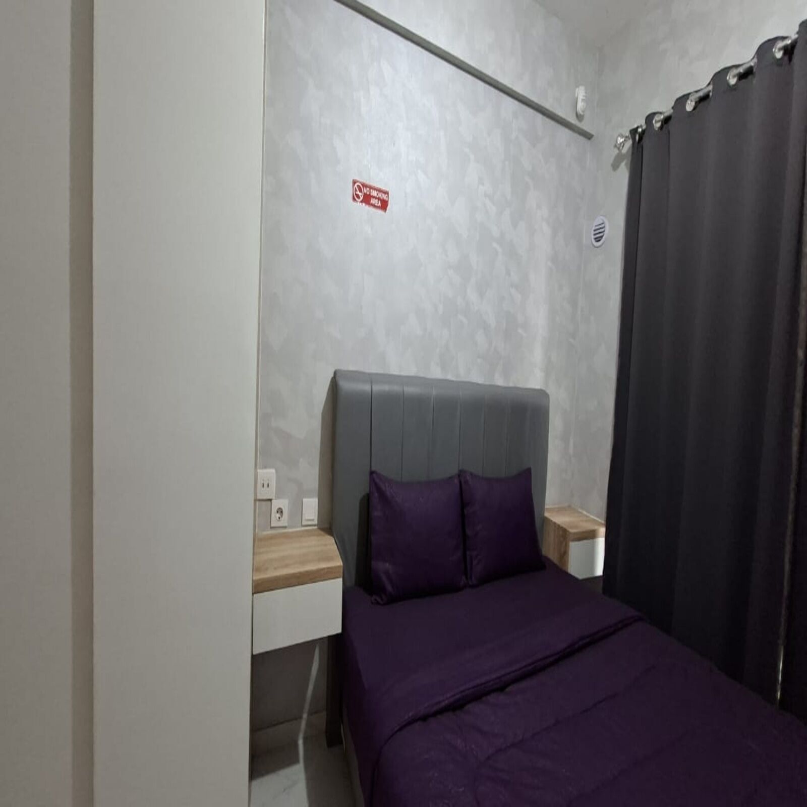 A bedroom with a bed, a nightstand, a power outlet, and a 'No Smoking' sign on the wall.