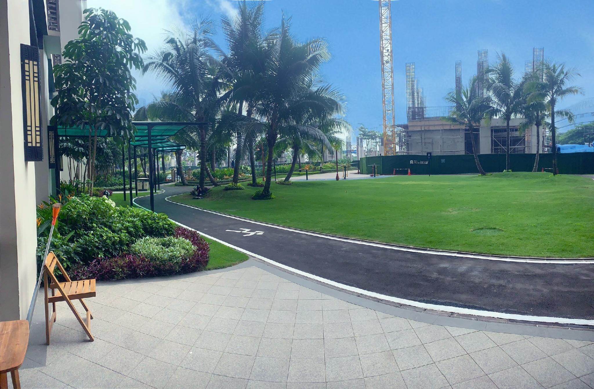 A paved pathway leading to a green lawn with palm trees, a construction site in the background, and a building with a green awning.