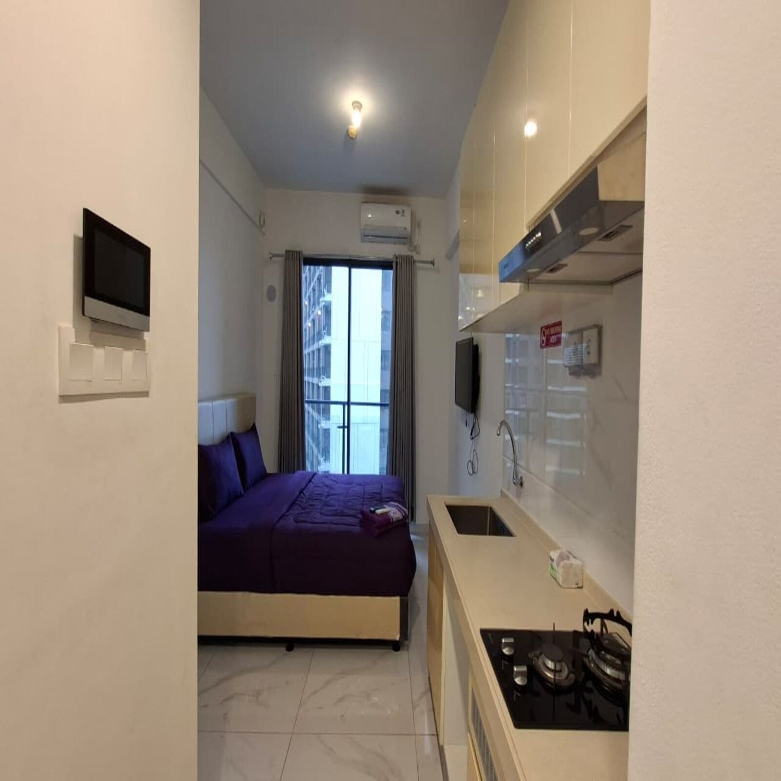 A compact room with a bed, a small kitchenette, a TV mounted on the wall, and a window with curtains.
