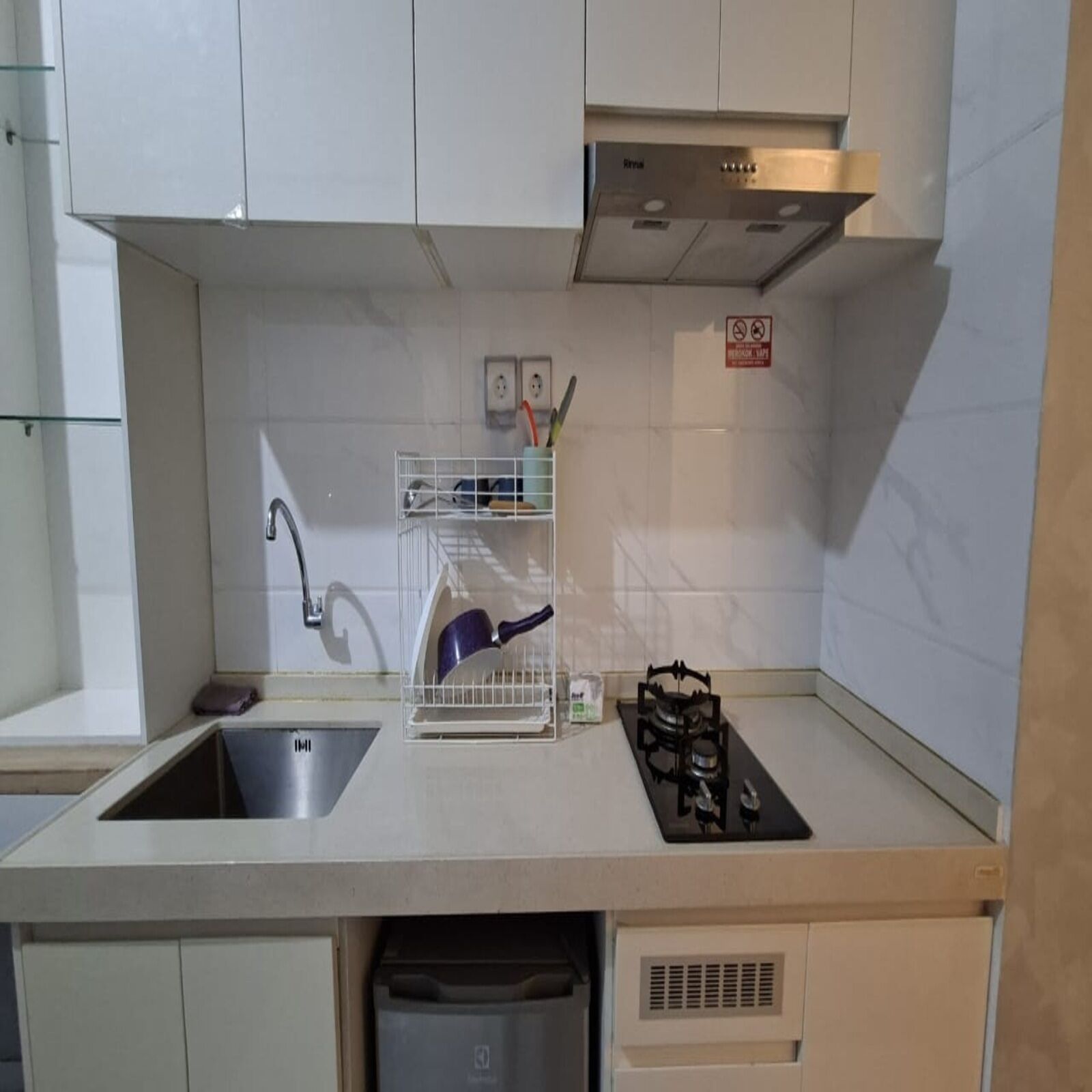 A compact kitchen with a sink, a dish rack, a stove, and a trash bin.