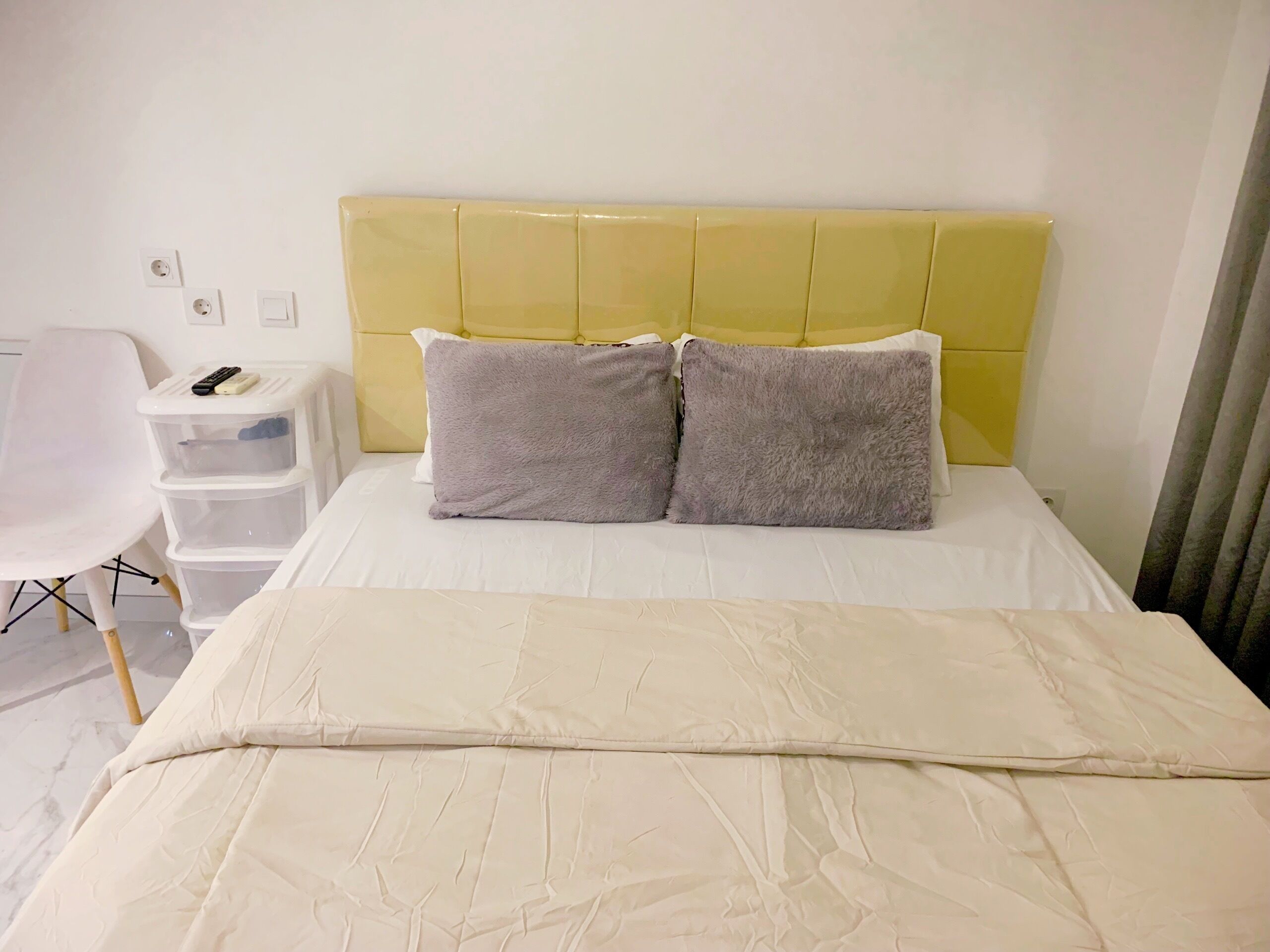 Een netjes opgemaakt bed met een witte deken, twee grijze kussens en een geel hoofdeinde. Er staat een witte stoel met een afstandsbediening erop, en een set witte opbergbakken tegen de muur.