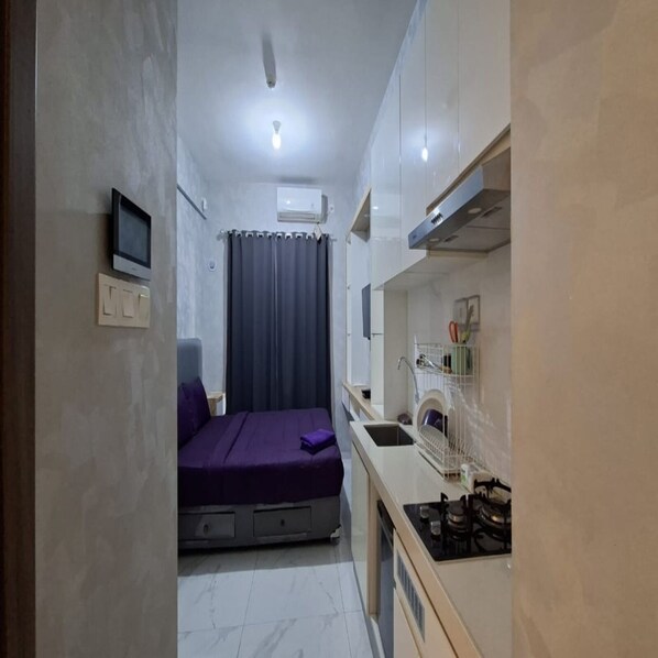 A compact room with a bed, a small kitchenette, a TV, and an air conditioning unit.