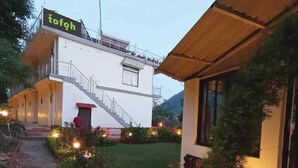 A two-story building with a staircase leading to the upper floor. There is a sign with the word "tofoh" on the roof. The building is surrounded by a well-maintained garden with various plants and a pathway. - Shiva solace by Sam Valley (Bhimtal)