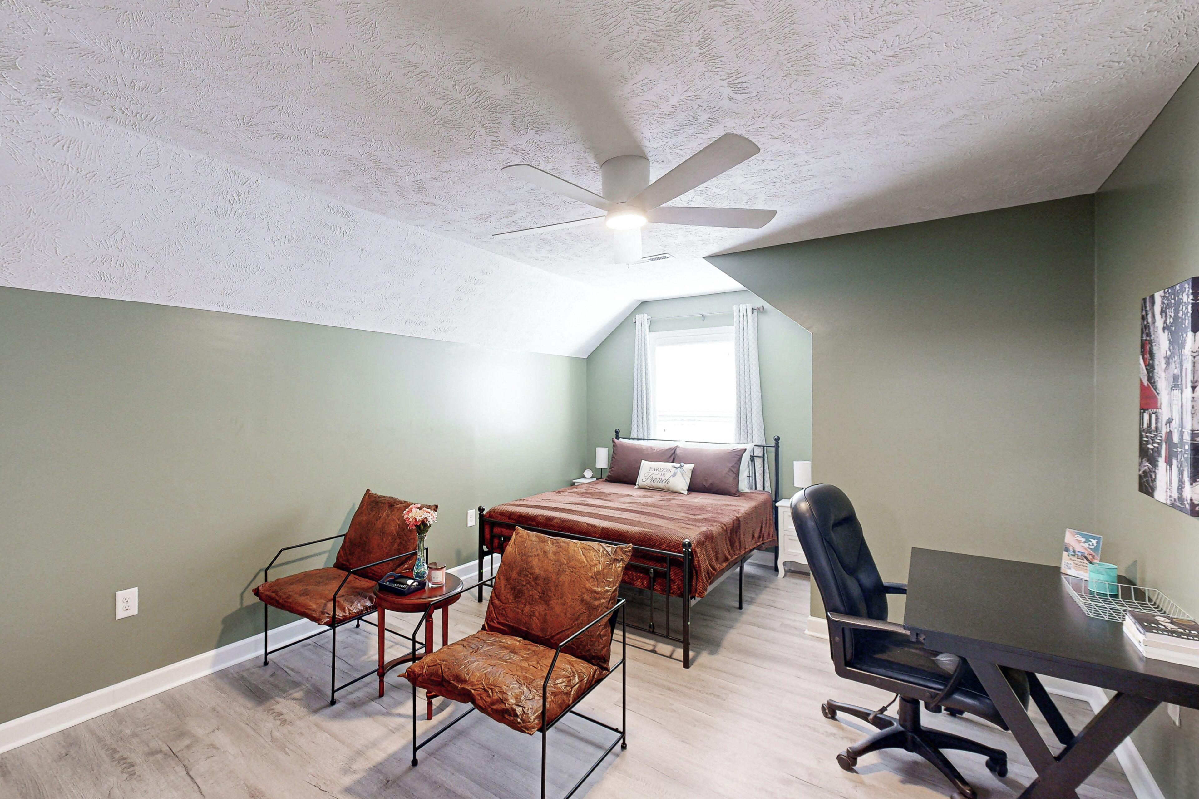 A bedroom with a bed, two chairs, a desk, and a ceiling fan.