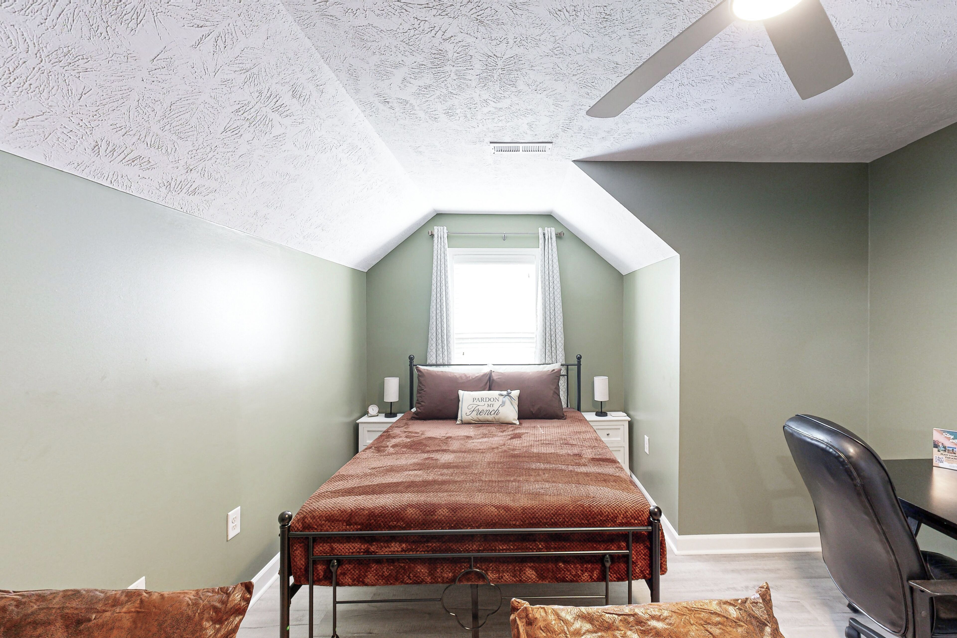 A bedroom with a sloped ceiling, a bed with a brown bedspread, a desk with a chair, and a window with curtains.