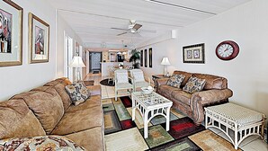 A spacious living room with a brown leather sofa, white armchairs, and a coffee table. There is a dining area in the background with a chandelier. - Shorefront Resort Retreat: Suntide III - Unit 1002 (South Padre Island)