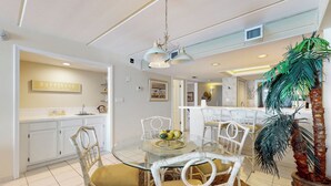 A dining area with a glass table, white wicker chairs, a central light fixture, and a kitchen in the background. - Shorefront Resort Retreat: Suntide III - Unit 1002 (South Padre Island)