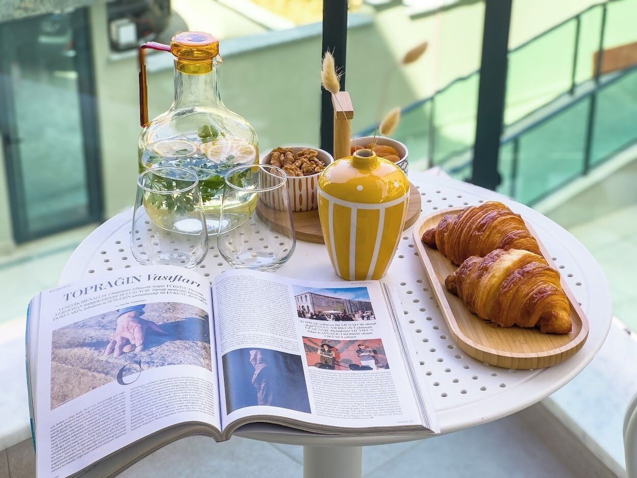 A table with a book, a bottle, a bowl of nuts, a yellow container, and a croissant on a tray.