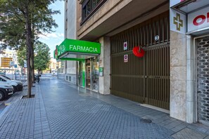 Uma farmácia com placa verde, entrada protegida por grades e calçada de azulejos.