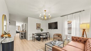 Een moderne woonkamer met een bank, salontafel en een eethoek met stoelen. Er hangt een televisie aan de muur en er is een kroonluchter.