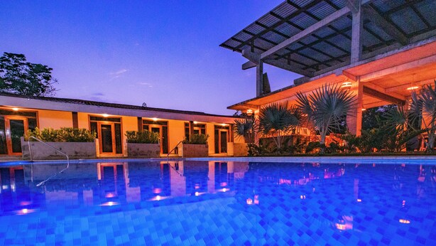 A swimming pool with a tiled floor, surrounded by a building with multiple doors and windows, and a covered area with plants. - Casa Las Flores (El Cuco)