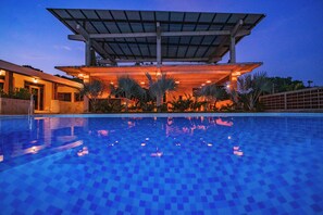 Piscina com padrão quadriculado, rodeada por palmeiras e uma área coberta com telhado.