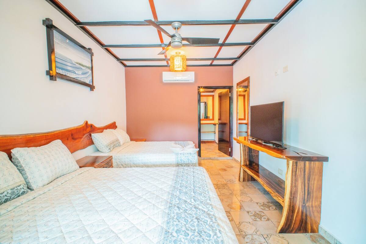 Un dormitorio con dos camas, cabeceros de madera, ventilador de techo, equipo de aire acondicionado, televisor sobre un soporte de madera y un cuadro en la pared.