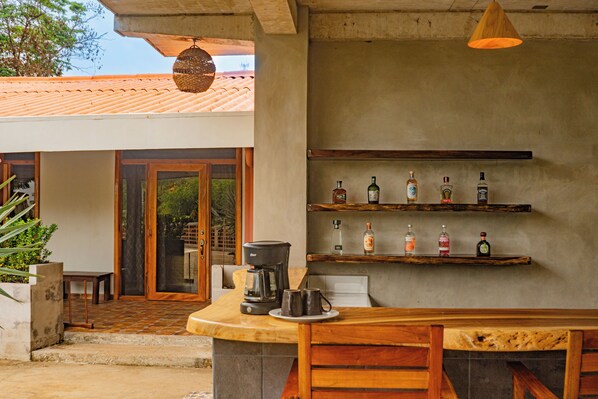 A kitchen area with a coffee maker, mugs, and a shelf with bottles. - Casa Las Flores (El Cuco)