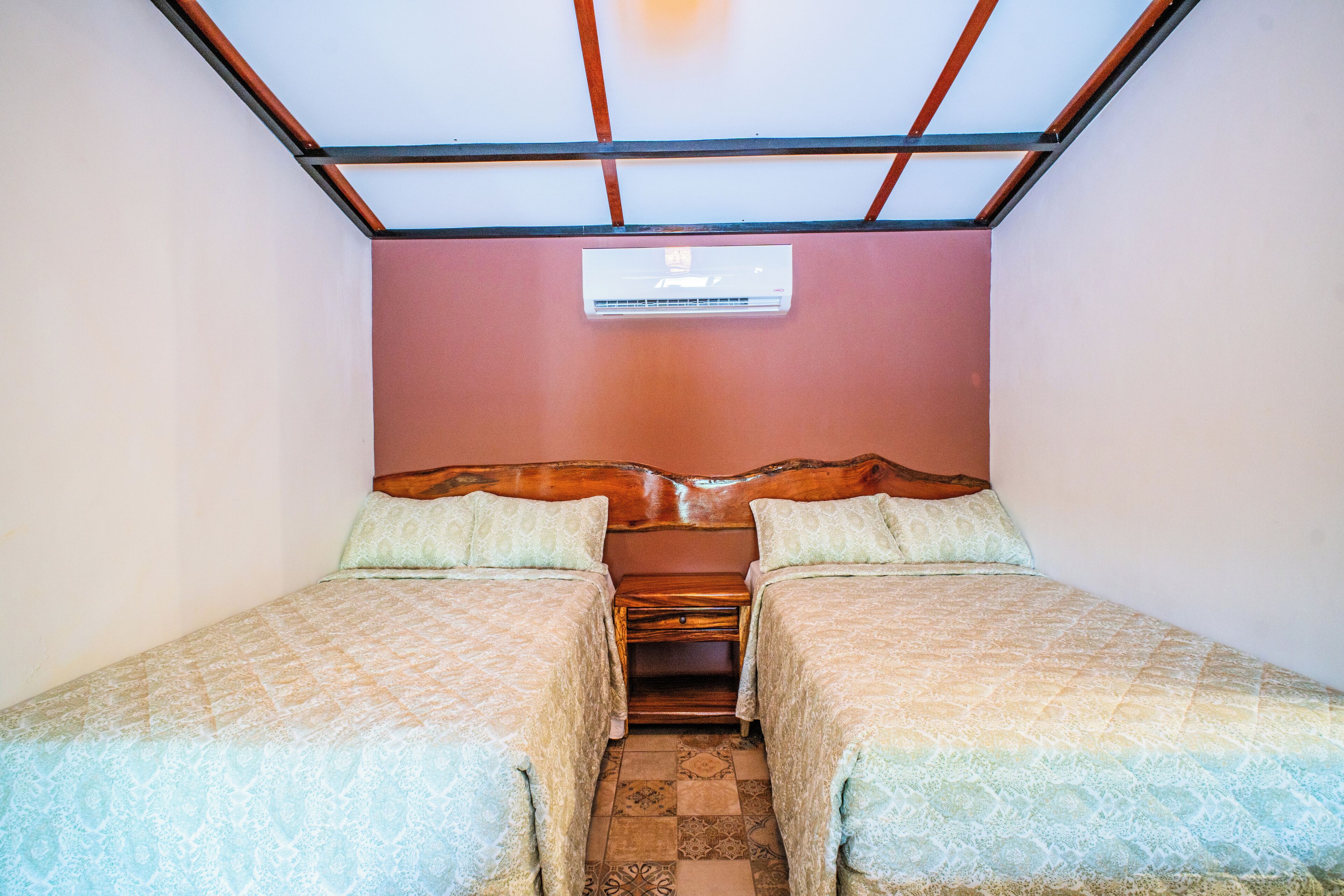 Une chambre avec deux lits simples, une tête de lit en bois, une petite table de chevet et un appareil de climatisation fixé au mur.