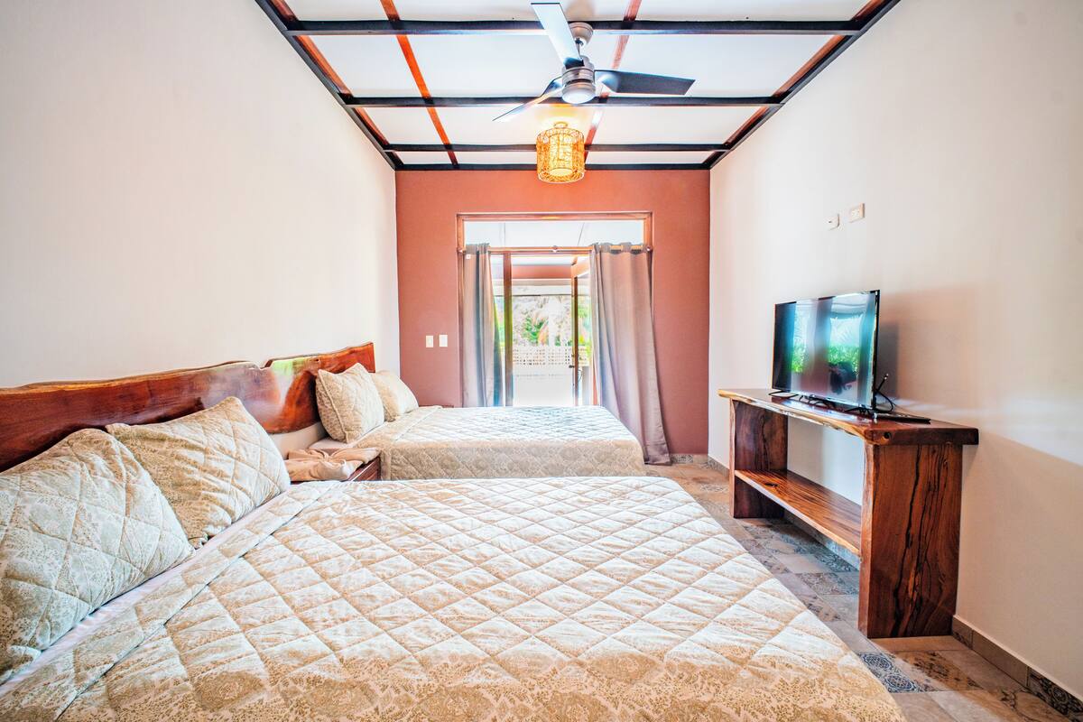 Un dormitorio con dos camas, un ventilador de techo, un televisor sobre un soporte de madera y vistas al exterior a través de una puerta.