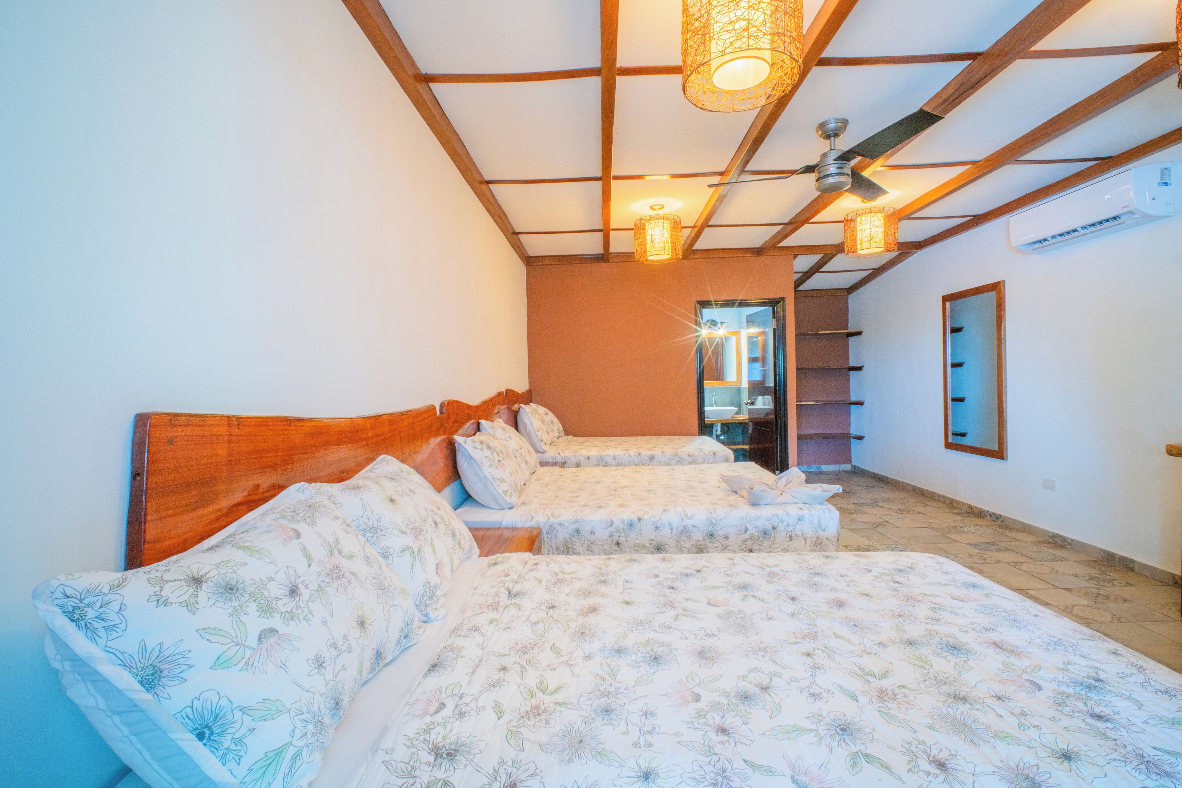 A bedroom with two beds, wooden headboards, a ceiling fan, and a hallway leading to a bathroom.