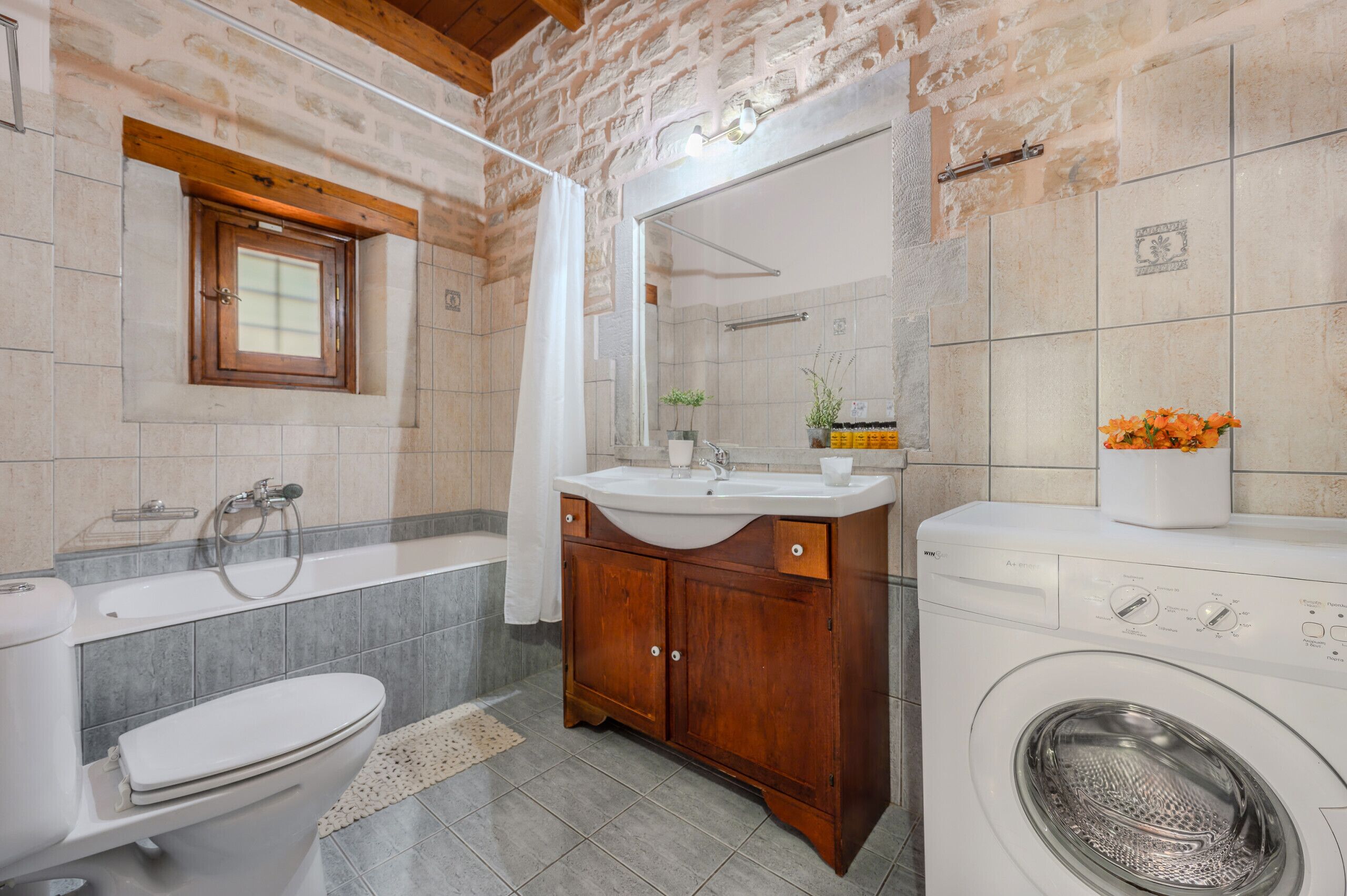 Une salle de bain avec un mur en pierre, un meuble en bois, un lavabo, des toilettes, une baignoire et un lave-linge.