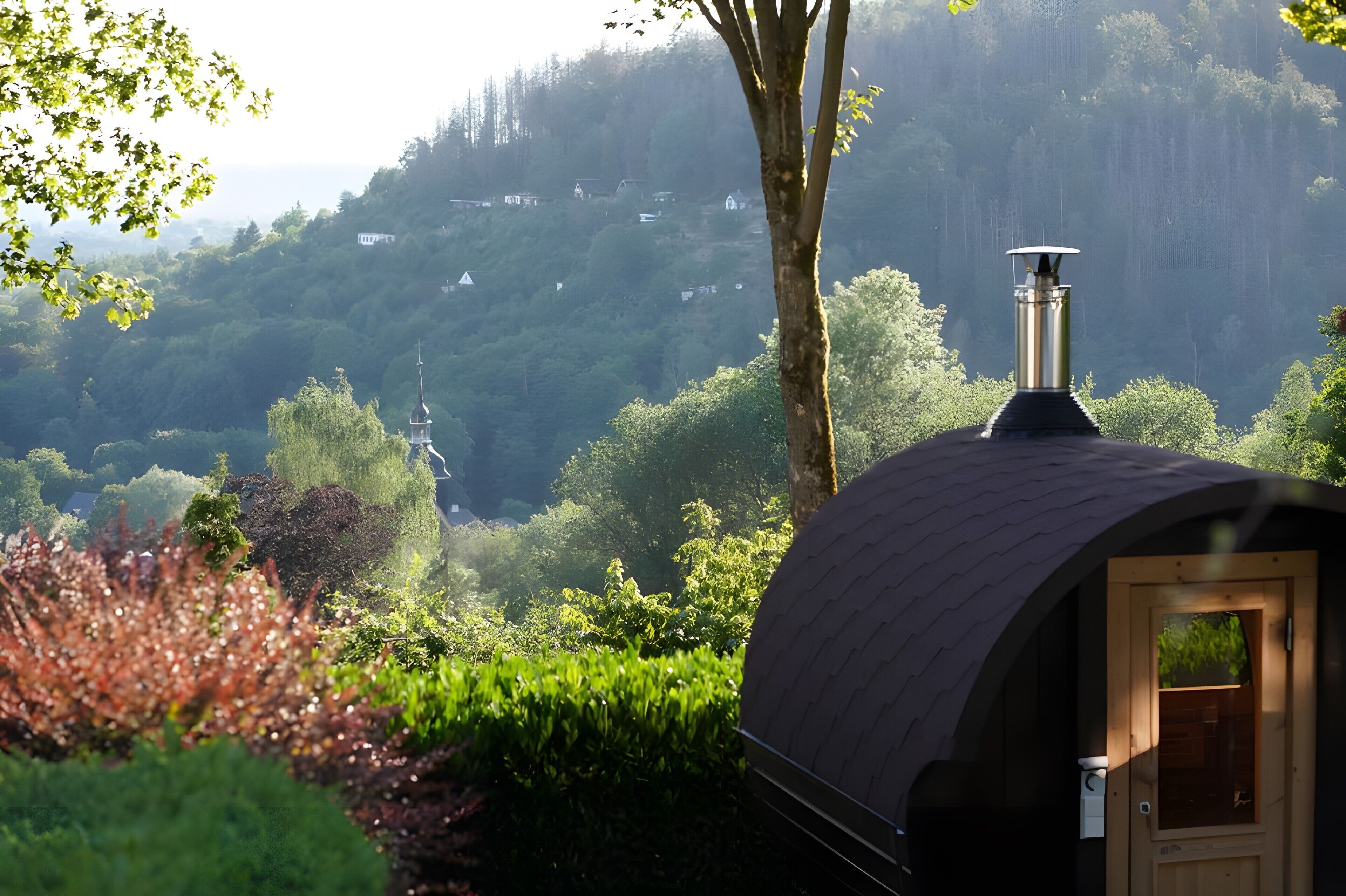 A dome-shaped building with a chimney, surrounded by lush greenery and a forested hillside in the background.