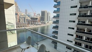 Ein Balkon mit zwei weißen Stühlen, einer Glastrennwand und Blick auf einen Fluss mit Gebäuden und Baukränen im Hintergrund.