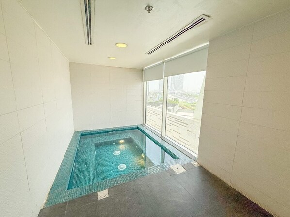 Una piscina interior pequeña con baldosas azules cristalinas, un ventanal con vistas a edificios e iluminación empotrada en el techo.