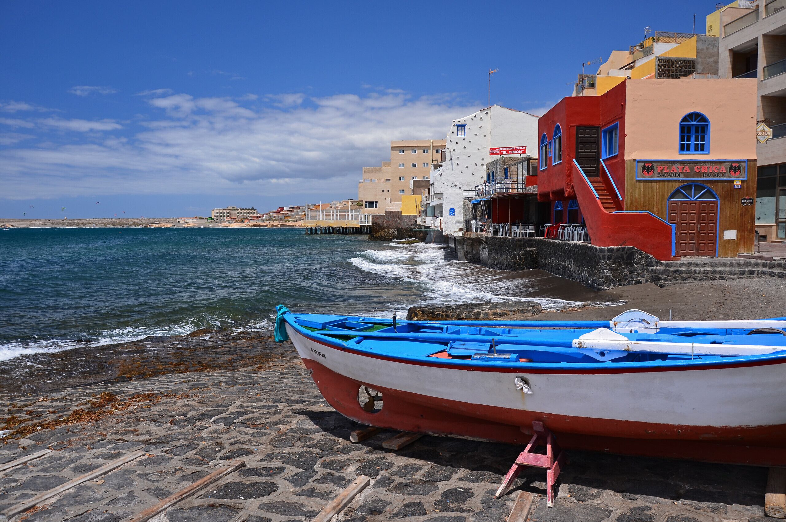 Escena costera con un bote en un muelle de piedra, edificios con letreros y un cielo despejado.