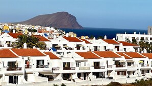 Uma cidade costeira com edifícios brancos, telhados de barro avermelhados e uma montanha imponente ao fundo.