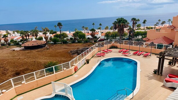 Piscina com design curvo, rodeada por uma cerca branca, com vista para uma praia com palmeiras e um céu claro.