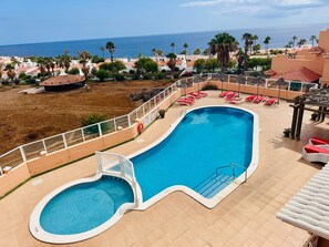 Une piscine au design incurvé, entourée d’une clôture blanche, et offrant une vue sur une plage bordée de palmiers et sous un ciel dégagé.