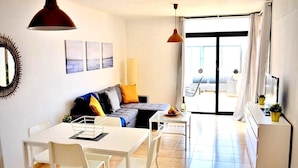Uma sala de estar moderna com sofá, mesa de jantar e cadeiras, área de cozinha com geladeira e uma grande janela com cortinas.