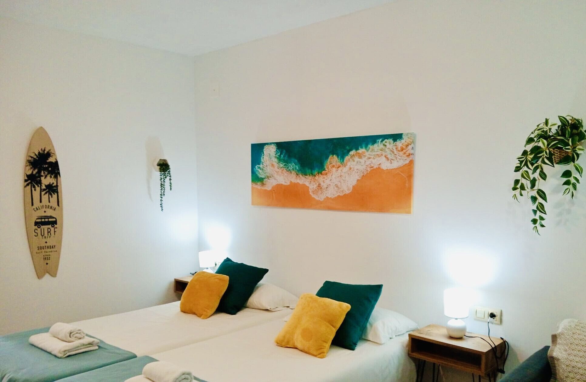 A hotel room with two beds, a surfboard decoration, a painting, and a hanging plant.