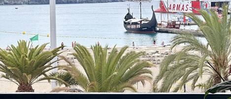 Vista costeira com um grande navio chamado 'ARMAS' ancorado no porto, palmeiras e um terraço com azulejos e cadeiras.
