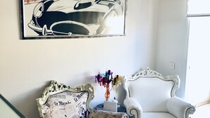 Una habitación con un cartel de coche antiguo, un sillón estampado y un sillón blanco clásico.