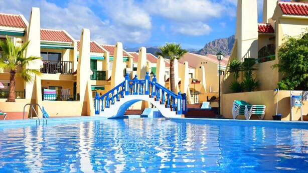 Una piscina con un elemento acuático azul, rodeada de edificios con techos de tejas rojas y balcones.