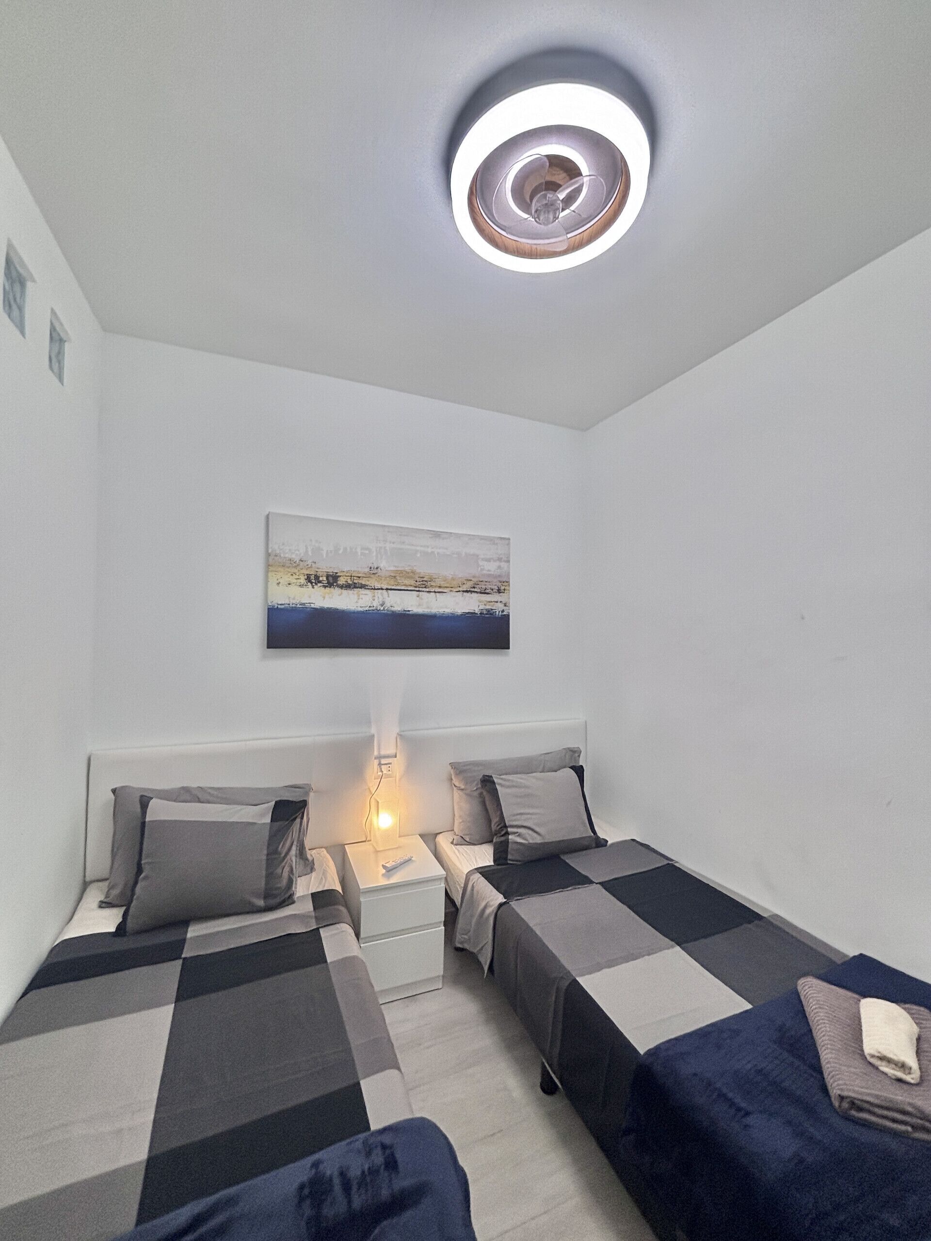 Una habitación pequeña y moderna con dos camas, una mesita de noche, una lámpara de pared y un cuadro.