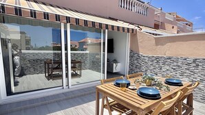 A modern outdoor dining area with a wooden table and chairs, a glass door leading to a patio, and a building with a striped awning in the background. - A revisar (Los Cristianos)