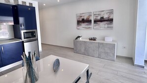 A modern kitchen with dark blue cabinets, a white countertop, and a dining area with a white table and blue chairs. There are two abstract paintings on the wall and a gray console table with decorative items. - A revisar (Los Cristianos)