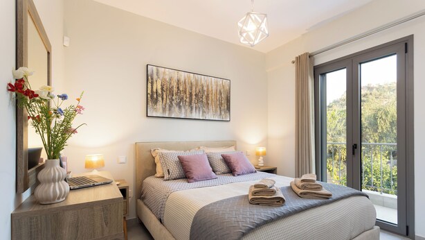 A modern bedroom with a large bed, a bedside table with a laptop, a vase of flowers, and a painting on the wall.