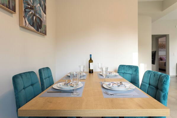 A dining table set with plates, glasses, and cutlery, flanked by teal upholstered chairs, with a bottle of wine on the table.
