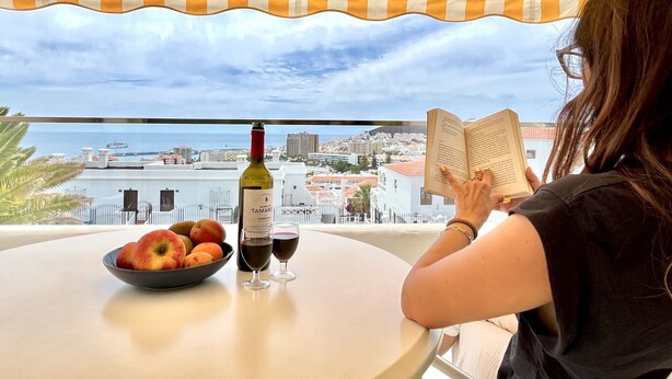 A person reading a book at a table with a bowl of fruit, a bottle of wine, and two glasses on a balcony with a view of the sea and cityscape.