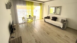 A modern living room with a sofa, a dining table with chairs, a TV, and a balcony with yellow curtains.