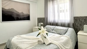 A modern hotel room with a neatly made bed, a painting of mountains, a window with sheer curtains, and a small bedside table with a tissue box.