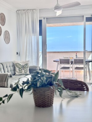 Uma sala de estar moderna com um sofá, uma mesa de jantar com cadeiras, um ventilador de teto e vista para o mar através de grandes janelas.
